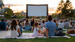 © AnastasiiaRozhmanova - Twilight outdoor cinema event with families on blankets facing inflatable screen in municipal park, warm dusk sky behind, children eating popcorn, adults reclined, summer evening community gathering