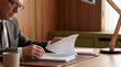 © Watercolor_Kawaii - Close up of senior businessman reading documents at wooden desk with cup and desk lamp, focused on paperwork in professional office environment