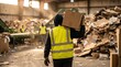 © Olga Ко - Ouvrier en gilet de haute visibilité portant une boîte en carton dans un grand entrepôt de recyclage ou une déchetterie