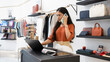 © fizkes - Indian woman stands at checkout counter of modern boutique, having business conversation on phone, working on laptop, check stock, managing online orders, handling customer service or logistics tasks