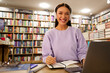 © Home-stock - Student wearing headphones writing notes and smiling at camera at library desk