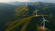 © Nasos Zovoilis - Wind turbines line green hills, harvesting wind for clean, renewable electricity; transmission lines and clear sky frame an energy transition landscape promoting sustainability and progress