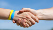 © pattozher - Solidarity Handshake: Two hands clasp in a firm handshake under a clear blue sky, one adorned with yellow and blue wristbands, symbolizing unity and support.