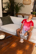 © wavebreak3 - Man wearing red jersey sitting on sofa at home holding smartphone, popcorn bowl, showing wristwatch