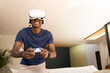 © wavebreak3 - African American man wearing white VR headset, holding controller, leaning over sofa at home