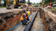 © MystoganCreative - Construction workers installing a large black drainage pipe in a deep trench on a sunny day.