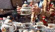 © Izi18 - Woman holding decorative porcelain teapot while examining vintage dishware at a flea market, surrounded by various antique items and collectibles on display