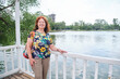 © Carolina Jaramillo - A senior hispanic woman smiles and looks at the camera as she enjoys her free time in a park by a lake. Concept: the well-being of older adults who stay active.