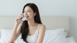 © CrazyJuke - A Refreshing Sip of Wellness: A young woman with long, dark hair smiles as she enjoys a cool glass of water, embodying health and hydration in a serene bedroom setting.