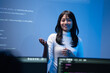 © Jestercine - Happy young Asian female programming instructor in a white turtleneck smiling and gesturing with her hand while teaching a coding class in front of a blue digital code wall