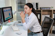 © MINAE - Young asian woman feeling stressed and overwhelmed by excessive workload, experiencing burnout and fatigue while sitting at her office desk with a headache