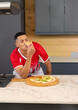 © wavebreak3 - Man wearing red jersey tasting pizza slice while leaning on marble countertop with wooden paddle