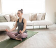 © wavebreak3 - Adult woman sitting cross-legged on green yoga mat in living room, wearing mauve leggings and watch