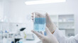© Watercolor_Kawaii - Scientist hands holding clear glass beaker with blue liquid in laboratory setting, wearing white gloves for safety and precision in research environment