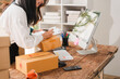 © Kritdanai - Small Business Owner Working: A dedicated female entrepreneur diligently organizes packages and writes on a clipboard, surrounded by her workspace filled with shipping boxes and a computer.