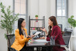© Kritdanai - Business Meeting in Office: Two businesswomen in formal attire engage in a discussion while seated at a desk in a modern office setting, surrounded by plants and office supplies.