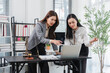 © Kritdanai - Professional Collaboration: Two business women intently review a project on a laptop in a modern office setting, engaged in focused discussion and problem-solving.