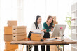 © Kritdanai - Small Business Owner Teamwork: Two Asian women, a small business owner and her employee, collaborate joyfully at a desk, surrounded by shipping boxes and organized shelves.