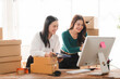 © Kritdanai - Small Business Owners Collaborating: Two smiling Asian women in a bright office space, surrounded by boxes, diligently work together at a computer.
