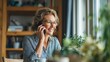 © Serhii - Smiling older woman on a phone call in a sunlit desk setup with plants and books, relaxed work-from-home scene