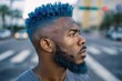 © ЮРИЙ ПОЗДНИКОВ - Profile view of a young African American man in his 20s with vibrant dyed blue hair and beard standing on a city street. He has dark skin and wears a grey shirt.