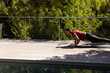 © wavebreak3 - Male performing plank on wooden deck beside pool wearing red shirt and dark pants, copy space
