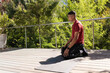 © wavebreak3 - Man kneeling on gray mat at balcony railing, wearing red shirt and showing wristwatch, copy space