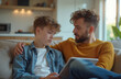 © miss irine - Father talks supportively to his sad teenage son at home on sofa. Young boy looks troubled with tablet screen while dad offers comfort and understanding during difficult conversation.