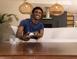 © wavebreak3 - African American man sitting on sofa in modern living room holding white controller smiling
