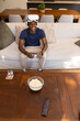 © wavebreak3 - African American man sitting on sofa in living room holding controller wearing white VR headset