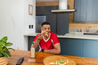 © wavebreak3 - African American man sitting at table in modern kitchen holding pizza slice wearing red shirt