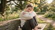 © Mateus - A young woman sits alone on a park bench, eyes closed, embracing herself in a moment of quiet contemplation. Sunlight filters through the trees, casting a warm glow on the serene scene