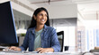 © fizkes - Cheerful Indian businesswoman sits at desk, working on pc, looks into distance with contented smile during workday in office. Workflow using modern device, internet resources, new AI digital tools