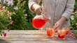 © Ivul - Enjoying a delightful summer afternoon in Canada, a refreshing red beverage is gracefully poured into chilled glasses on a rustic wooden table amidst blooming garden flowers