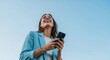 © Hungarian - Happy Young Businesswoman Laughing While Using Her Smartphone Outdoors Against a Clear Blue Sky Background
