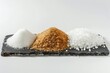 © TobiStocks - A trio of sweet crystals: fine white sugar, coarse brown sugar, and rough white sugar granules are presented on a slate serving platter, inviting exploration of sweetness.