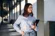 © standret - Young woman with brown hair wearing a light blue shirt and jeans stands confidently in a modern corridor, holding a tablet and smiling softly