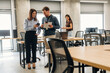 © standret - Two professionals discussing project details in a modern office space with desks and computers, while a woman works on a laptop in the background