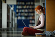 © TrueFrame Collective - Female student studying on laptop while sitting on library floor, focusing on online learning and academic research