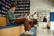 © TrueFrame Collective - Male student studying on laptop in library, wearing headphones and preparing coursework while classmates learning nearby. Useful for education marketing, e learning, student life