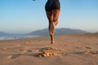 © sutulastock - Back view close-up of athletic woman jogging barefoot on beach at sunrise, sand flying from her foot with every step, warm morning light and soft mountain horizon in background.