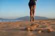 © sutulastock - Back view of a fit barefoot woman running on sand at sunrise, kicking up sand with each step, showcasing toned legs, strength, and motion in a scenic coastal landscape.