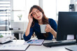 © Fahng - Stressed business woman rubs her temple while holding a coffee cup and working on a laptop in office, fatigue, frustration and  stress.