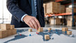 © vatcharapong - supply chain management logistics risk management. Businessman placing wooden blocks on a world map in a warehouse, symbolizing global logistics and supply chain management.