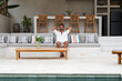 © BUDDHA - Man relaxing on a sofa in a modern outdoor living area with a pool