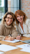 © pattozher - Two confident women collaborating at a desk, smiling and looking directly at the camera, conveying warmth and professionalism.