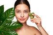 © Oleg Gekman - Close up woman face with green exotic leaf. Model doing skin care routine, scrapping, massage facial skin with jade gua sha scrapper