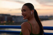 © Home-stock - Smiling woman in blue sports bra and wireless earbuds standing on bridge
