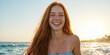 © Aleksei Solovev - Slender red-haired woman in blue strapless top, smiling at the camera, with wavy ocean and sunset in the background, hair blowing in the wind.
