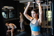 © JackF - Slender athletic girl work out muscles of arms and shoulders on simulator in gym of fitness club. Gym visitor trains triceps during classes at multi-station in wellness center.